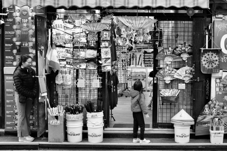 A young girl looking into a retail shop, while an older woman is hiding behind a door.