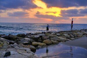Man standing on rocks, looking at the sun setting across the Mediterranean sea. 2020. © Amha Mogus